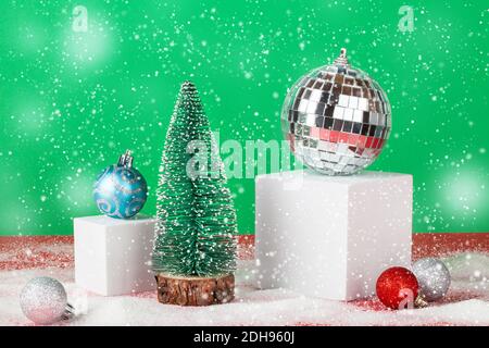 Weihnachten und Neujahr Komposition.Disco Ball und Kugel Dekoration auf weißem Würfelsockel mit Weihnachtsbaum auf rotem und grünem Hintergrund mit Schnee. Stockfoto