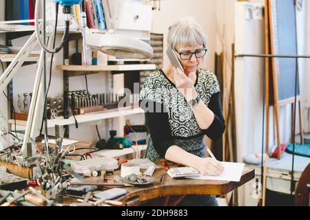 Ältere Frau mit Handy, während auf Dokument in Schmuck schreiben Werkstatt Stockfoto