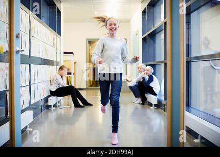 Lächelndes Mädchen läuft im Schulkorridor mit Freunden im Hintergrund Stockfoto