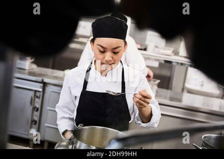 Junge weibliche Köchin, die Essen aus der Kochpfanne bei Commercial verkostet Küche Stockfoto