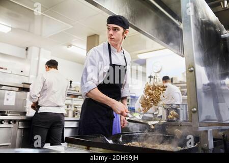 Männlicher Koch Student werfen Fleisch mit Kollegen im Hintergrund an Kochschule Stockfoto