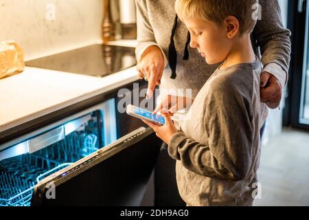 Junge lernt von der Mutter, während sie die mobile App über den Geschirrspüler nutzt In der Küche im Smart Home Stockfoto