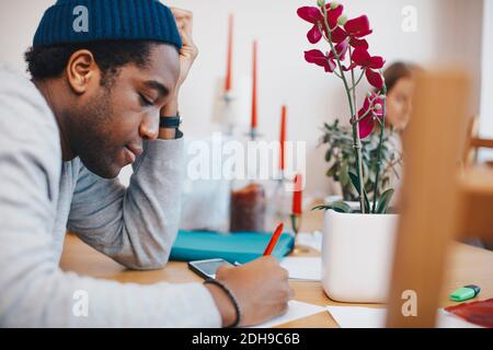 Seitenansicht des Mannes, der auf Papier am Tisch schreibt studentenwohnheim Stockfoto