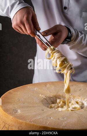 Im Restaurant bereitet ein Koch in blauer Kleidung Pasta Fettuccine Alfredo zu. Mischt Pasta in den Kopf von Parmesan. Der Hintergrund ist dunkel. Stockfoto