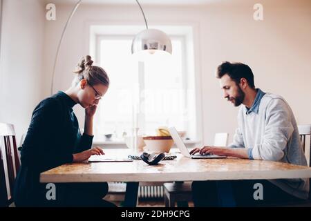 Seitenansicht des Paares mit Technologien am Esstisch während Arbeiten von zu Hause aus Stockfoto