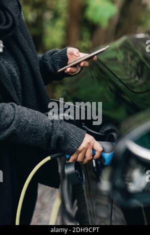 Mittelteil der mittleren erwachsenen Frau, die während des Ladevorgangs ein Mobiltelefon verwendet Elektroauto Stockfoto