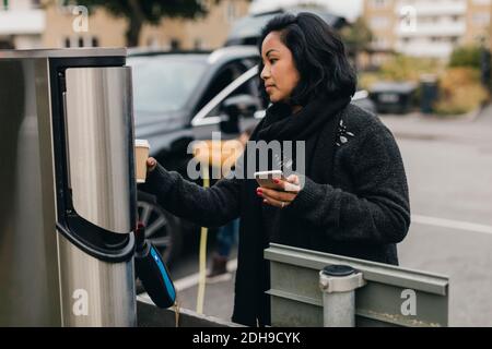 Mittlere Erwachsene Frau mit Smartphone beim Aufladen des Elektroautos Station Stockfoto