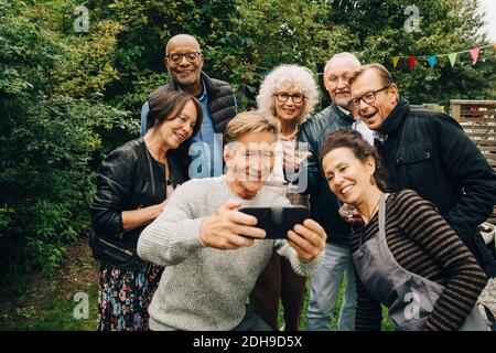 Lächelnder älterer Mann nimmt Selfie mit weiblichen und männlichen Freunden Während Gartenparty im Hinterhof Stockfoto