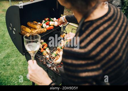 Mittelteil der Frau, die einen Drink hält, während sie das Abendessen auf der Rückseite grillen Hof Stockfoto