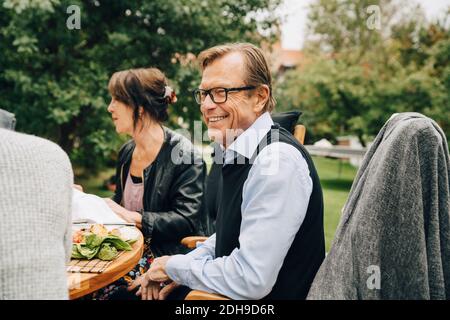 Lächelnder älterer Mann, der mit Freunden am Esstisch sitzt Hinterhof Stockfoto