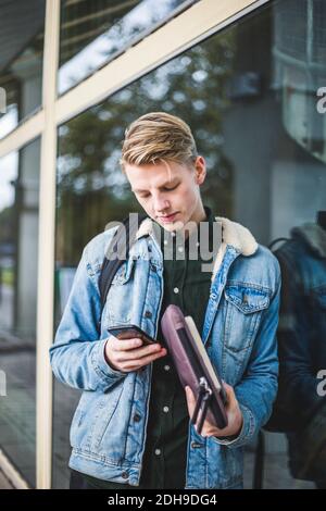 Junger Mann mit Handy, während er sich an einer Glaswand lehnte Stockfoto