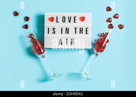 Festlicher Hintergrund mit leuchtendem Leuchtkasten mit Text Love is in the Air und Sektgläser mit roten Herzen auf blauem Hintergrund zum Valentinstag. Fl Stockfoto