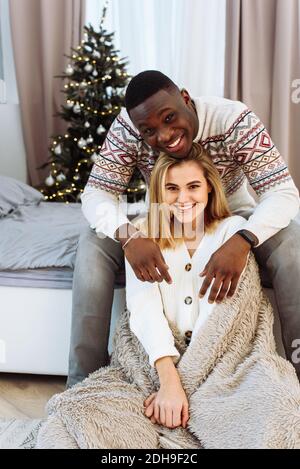 Junges glückliches Paar umarmt und entspannt auf einem bequemen Bett. Schönes multiethnisches Paar trägt weihnachtspullover und umarmt. Afrikanischer Mann und affecti Stockfoto