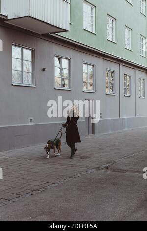 Die ganze Länge der jungen Frau, die mit Hund auf dem Fußweg läuft Durch Gebäude in der Stadt Stockfoto