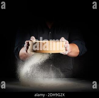 Koch ein Mann in schwarzer Uniform hält eine Runde Holzsieb in den Händen und siebt Weizenmehl Stockfoto