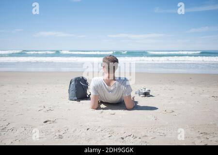 Rückansicht des Mannes, der mit dem Rucksack am Strand relaxt Sonniger Tag Stockfoto