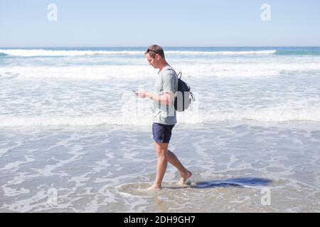 Volle Länge Seitenansicht von reifen Mann mit Handy Beim Spaziergang im Meer am Strand Stockfoto