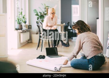 Frau, die auf dem Boden sitzt und mit einer Kollegin über die Blaupause diskutiert Zu Hause Stockfoto