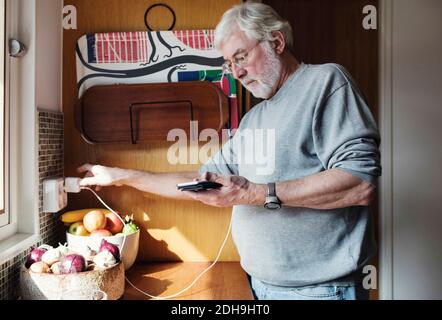 Senior Mann hält Handy, während das Ladegerät an die Stromversorgung angeschlossen wird Steckdose in der heimischen Küche Stockfoto