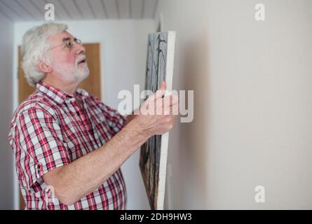 Älterer Mann hält Gemälde und schaut auf weiße Wand innen Wohnzimmer Stockfoto