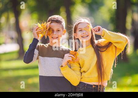 Glücklich Zwillinge Jugendliche junge und Mädchen posiert umarmt einander Im Herbst Park hält gefallene gelbe Blätter in der Hand Sonnig Stockfoto
