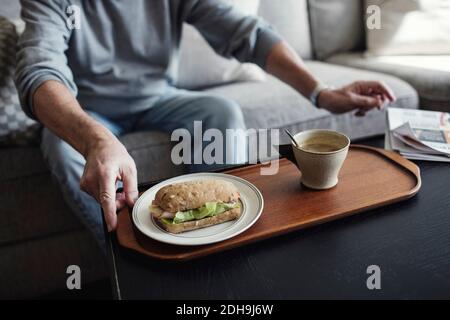 Mittelteil des älteren Mannes mit Sandwich und Tasse sitzen auf Sofa zu Hause Stockfoto