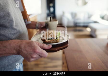 Mittelteil des älteren Mannes, der Tablett mit Sandwich und Tasse hält Zu Hause Stockfoto