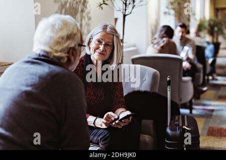 Ältere Frau mit Smartphone schaut beim Sitzen auf den Mann Auf dem Stuhl im Hotel Stockfoto