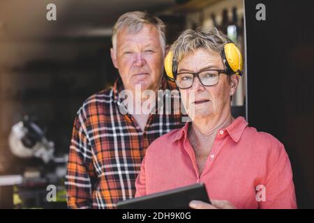 Porträt einer selbstbewussten älteren Frau, die im Stehen Ohrenschützer trägt Mit dem Mann an der Tür der Werkstatt Stockfoto