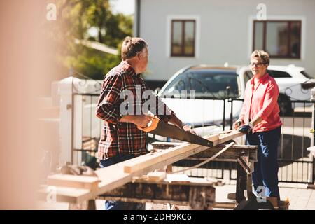 Älterer Mann, der die Handsäge hält, während er die Frau anschaut Holzbohlen im Hof Stockfoto