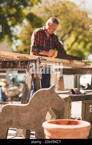 Low-Winkel-Ansicht von Senior Mann Schneiden Holzdiele mit Handsäge auf dem Hof Stockfoto