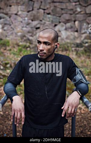 Nachdenklicher männlicher Athlet, der Musik hört, während er sich auf parallele Balken stützt Im Wald Stockfoto