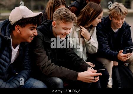 Lächelnde Mädchen und Jungen im Teenageralter, die während des Sitzens Mobiltelefone benutzen Im Park Stockfoto