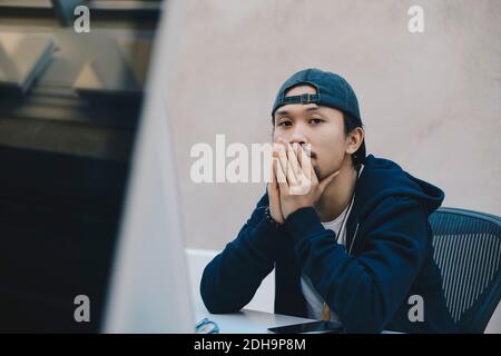 Männlicher Computerprogrammierer, der mit Händen sitzt, die den Mund am Computer bedecken Schreibtisch im Büro Stockfoto