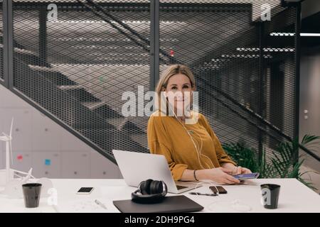 Porträt einer selbstbewussten Geschäftsfrau, die Kopfhörer trägt, während sie die Technologie bei nutzt Schreibtisch im Kreativbüro Stockfoto