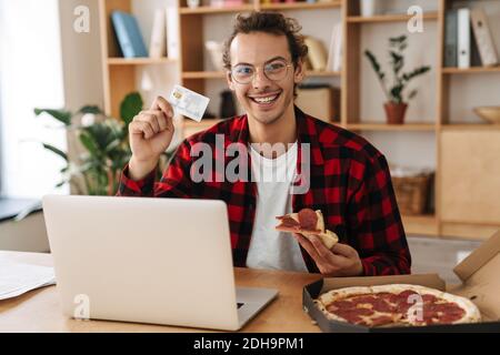 Schöner fröhlicher Kerl hält Pizza beim Essen Pizza und arbeiten Mit Laptop im Büro Stockfoto