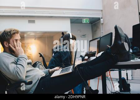 Müder Computerprogrammierer sitzt mit den Füßen auf dem Schreibtisch, während Kollegen, die im Hintergrund arbeiten Stockfoto