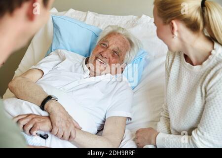 Glücklicher älterer Mann, der sich mit einem Paar unterhielt, während er sich auf dem Bett lehnend Auf der Krankenstation Stockfoto