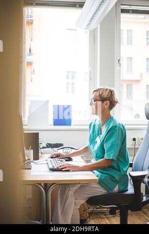 Seitenansicht des reiferen Arztes, der während des Sitzens über dem Computer arbeitet In der Klinik Stockfoto