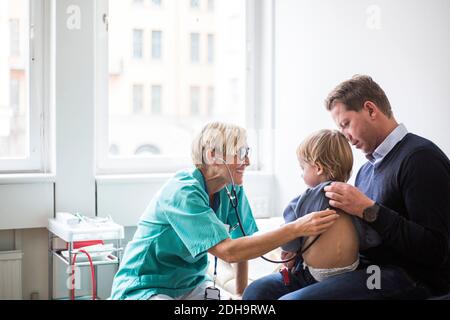 Lächelnde Ärztin, die den Rücken des Jungen mit Stethoskop untersucht, während sie sitzt Vom Vater im Krankenhaus Stockfoto