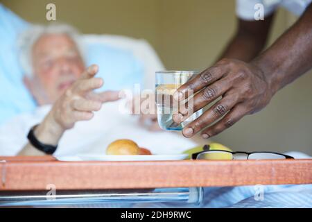 Zugeschnittenes Bild eines Krankenpflegers, der Senior mit Trinkglas übergab Mann auf der Krankenstation Stockfoto