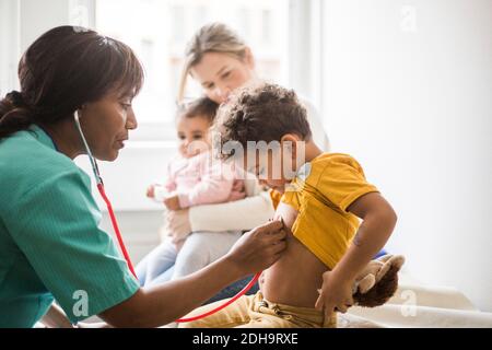 Seitenansicht einer Ärztin, die den Herzschlag des Jungen mit Stethoskop untersucht Während Mutter sitzt und Tochter in der Klinik Stockfoto