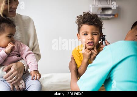 Portrait des Jungen, der Gesichter während der Untersuchung des Ohrs durch Frau macht Arzt in der Klinik Stockfoto