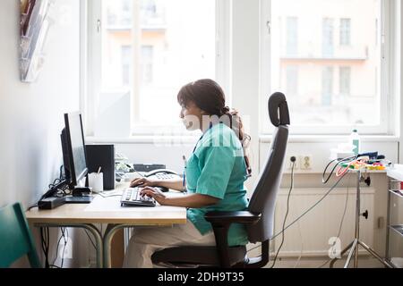 Seitenansicht der reifen weiblichen Arzt arbeiten über Computer, während Sitzen in der Klinik Stockfoto