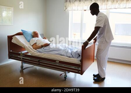 Krankenschwester, die das Bett anpasst, während ein älterer Mann Fernsehen schaut Krankenhausstation Stockfoto