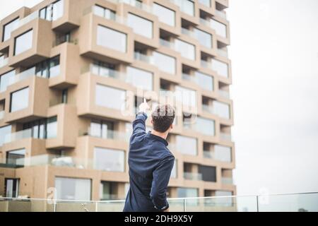 Rückansicht des Mannes, der beim Vorbeistehen auf das Gebäude zeigt Glasgeländer auf der Terrasse Stockfoto