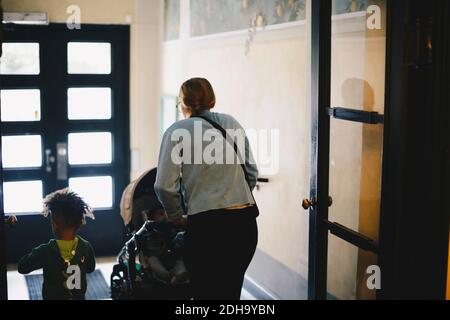 Rückansicht der Mutter, die den Kinderwagen beim Gehen mit schiebt Sohn an der Tür Stockfoto