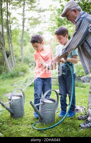 Großvater mit Enkeln hält Gartenschlauch beim Befüllen von Gießkannen Im Hinterhof Stockfoto