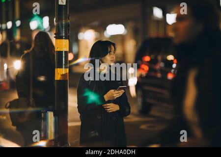 Junge Frau schaut weg, während Sie Handy in der Stadt an Nacht Stockfoto