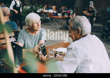 Ältere Frau, die dem Mann Smartphone zeigt, während sie ansitzt Restaurant in der Stadt Stockfoto